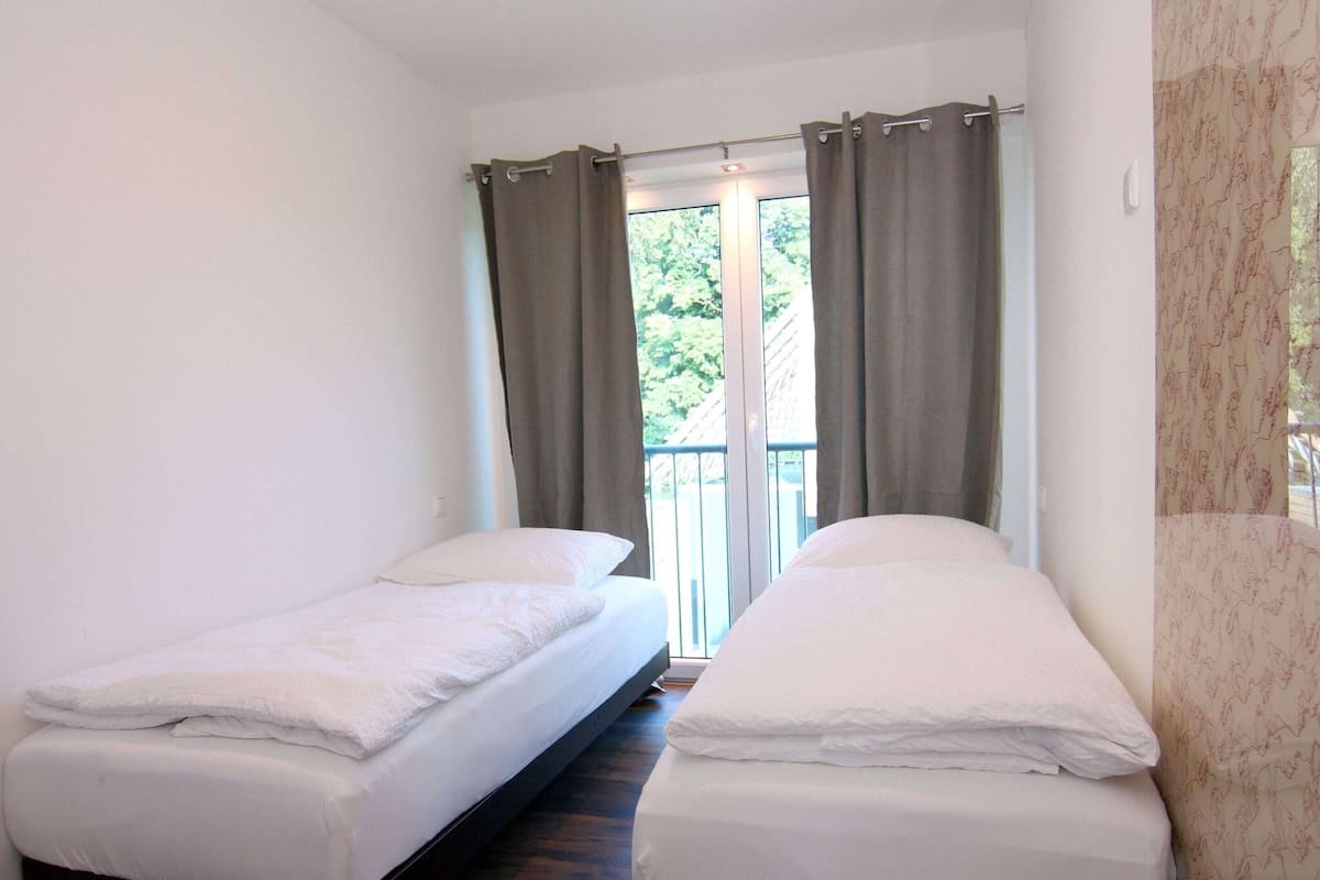 A room with two single beds, a window with curtains, and a view of greenery outside.
