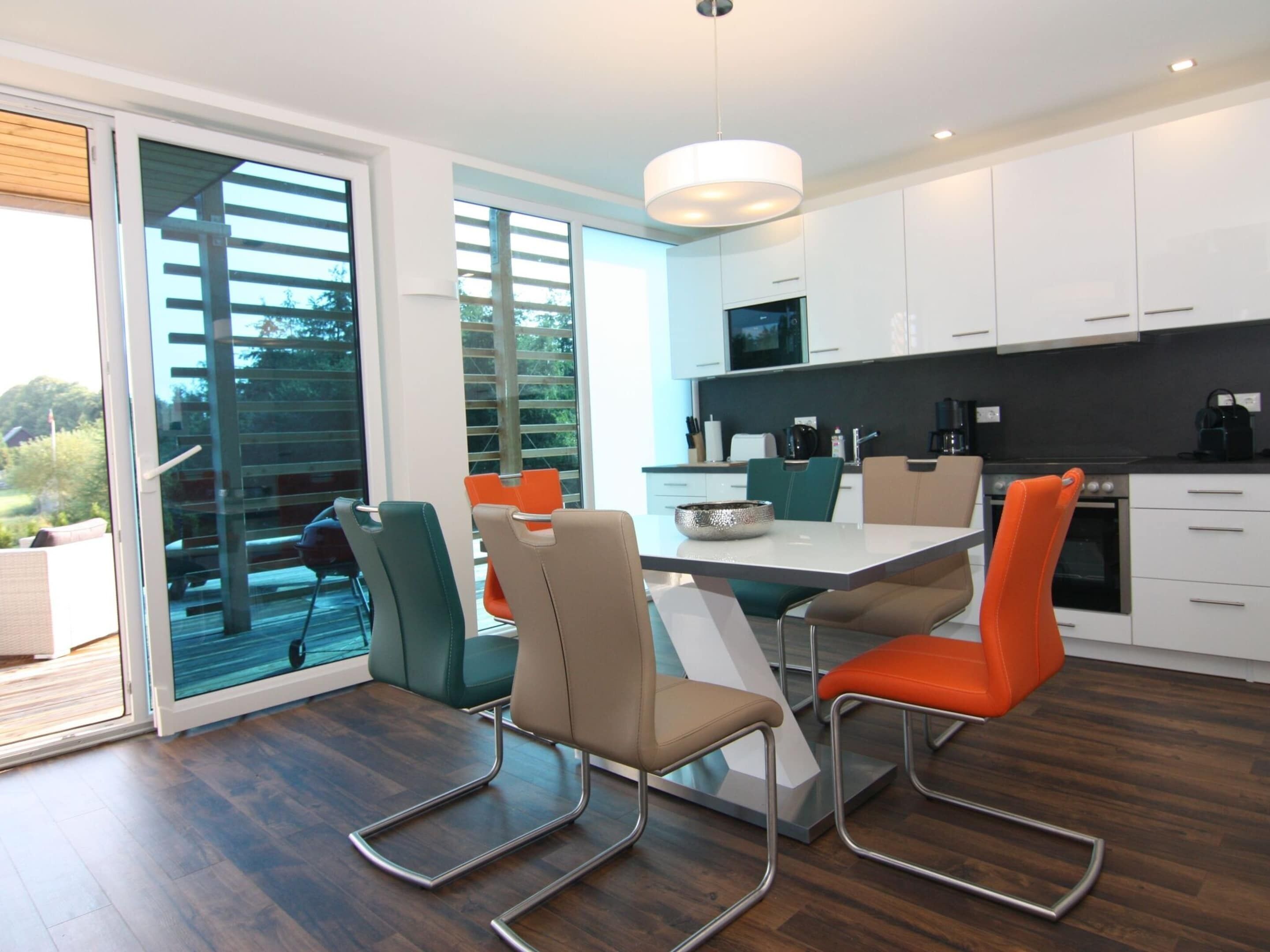 A modern kitchen with white cabinets, a black countertop, and a stainless steel oven. There is a dining table with four chairs, two of which are orange.