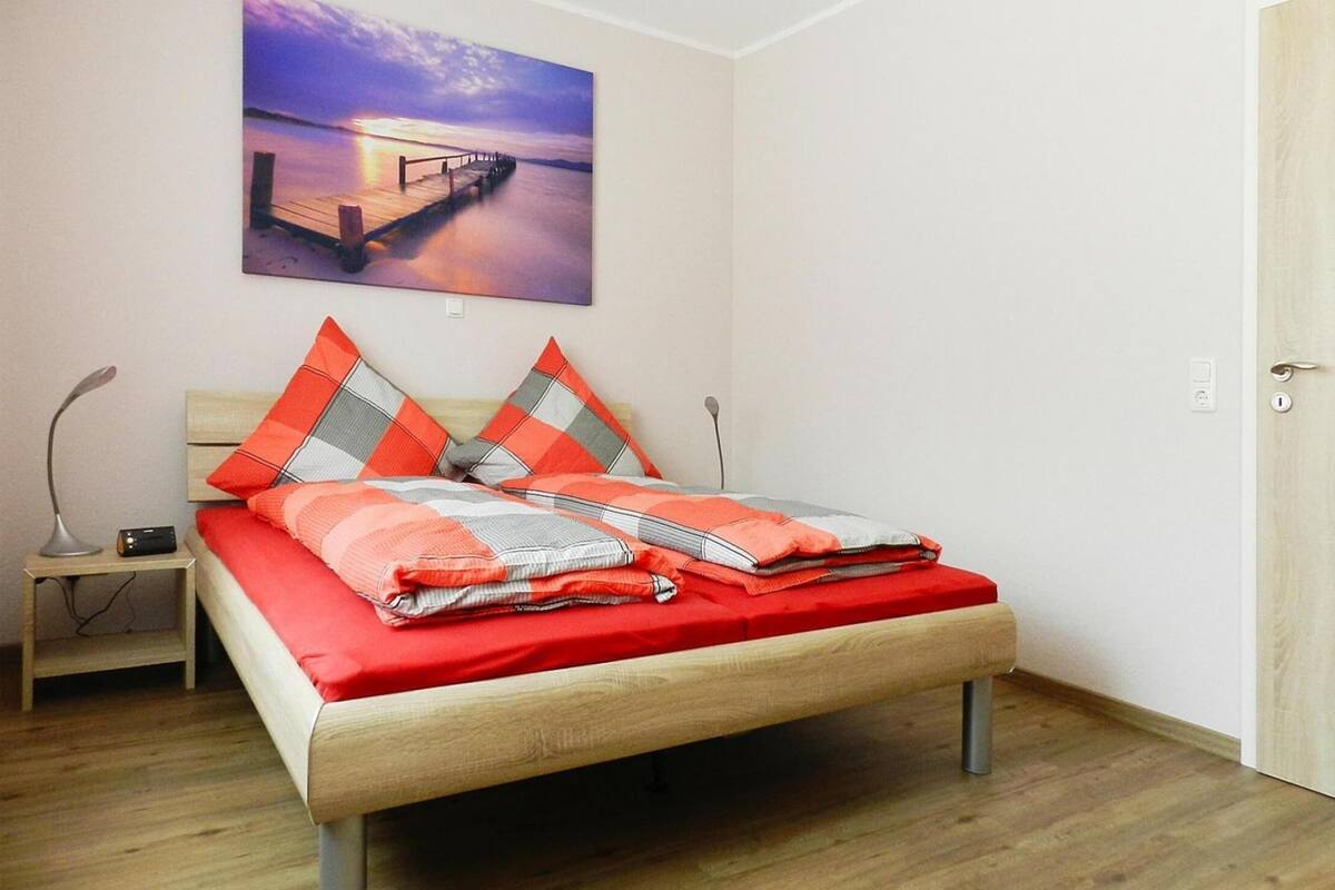A bedroom with a wooden bed, red and grey bedding, a nightstand with a lamp, and a painting of a pier on the wall.