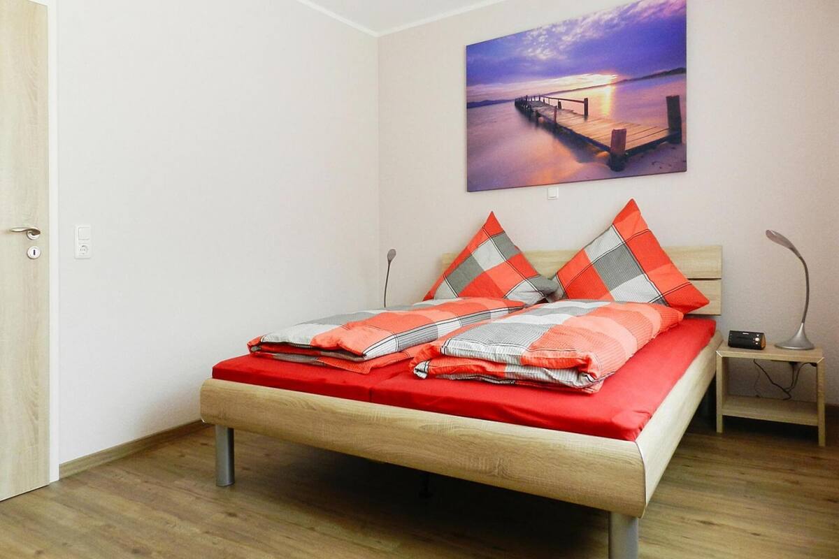 A neatly made bed with a red and grey checkered blanket, a wooden headboard, and two pillows with a red and grey checkered pattern. There is a nightstand with a lamp and a small device on it. A painting of a pier at sunset is on the wall.