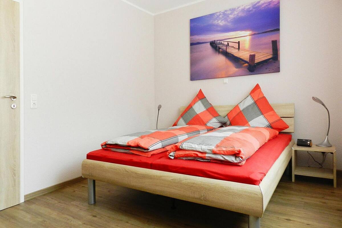 A bedroom with a wooden bed, a nightstand, a lamp, and a painting of a sunset over a pier.