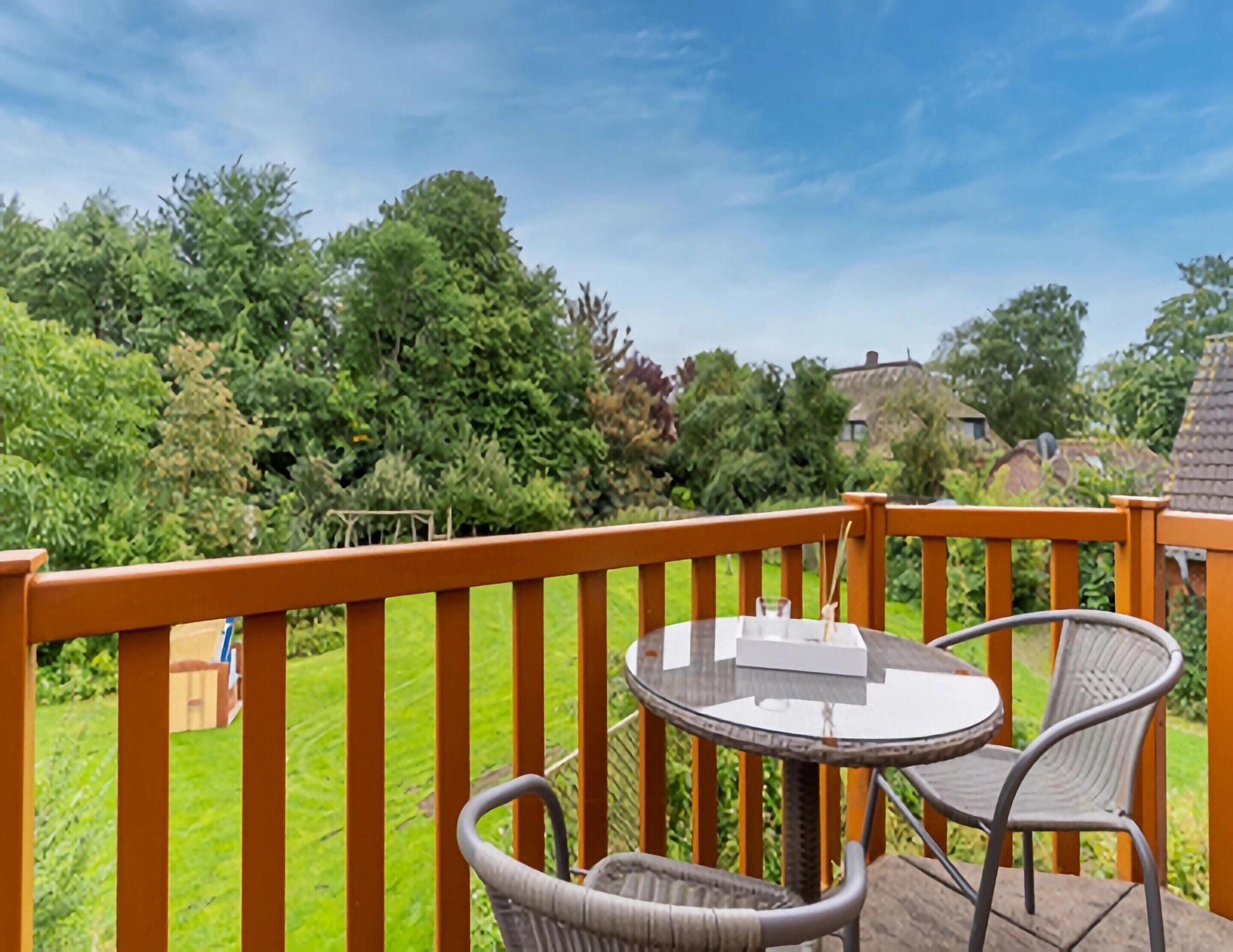 A balcony with a table and chairs, overlooking a garden with various trees and a house in the background.