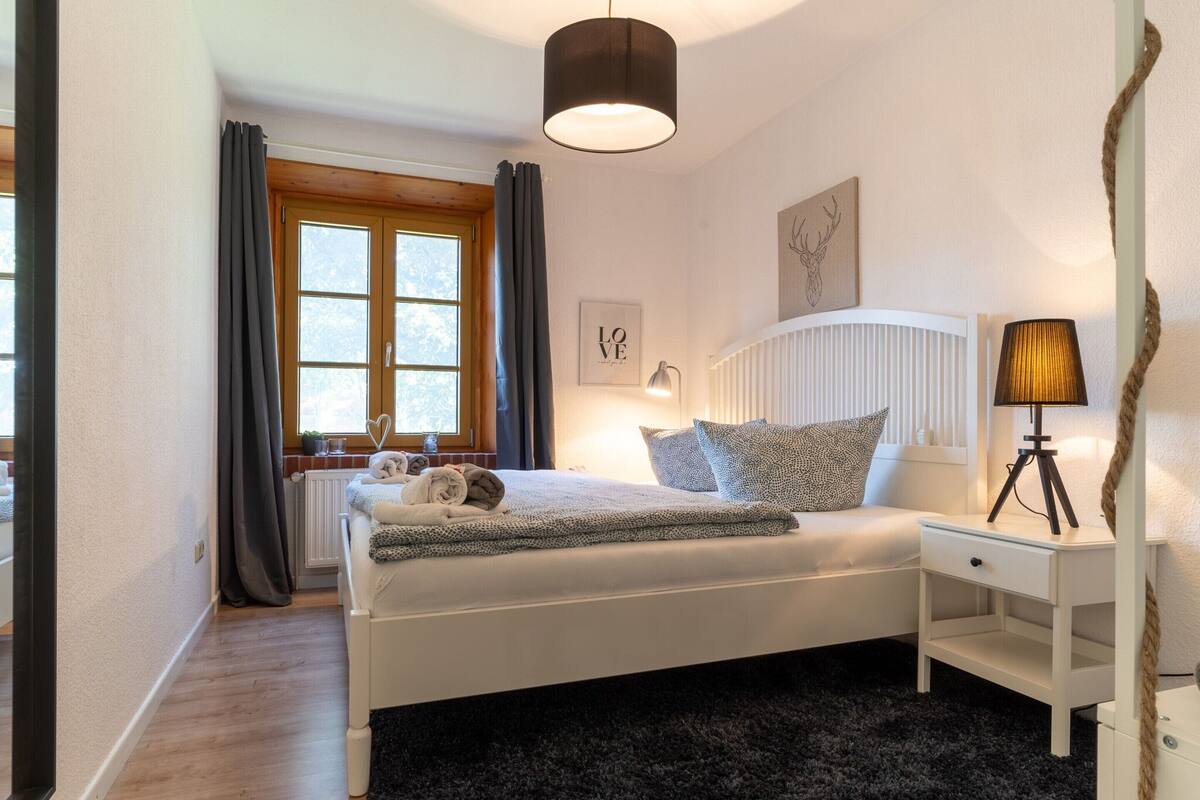 Une chambre à coucher comprenant un lit, une table de chevet, une lampe, une fenêtre et une œuvre d’art.