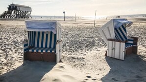 Deux chaises de plage avec des coussins à rayures, posées sur une plage de sable, une jetée en arrière-plan et un ciel dégagé.