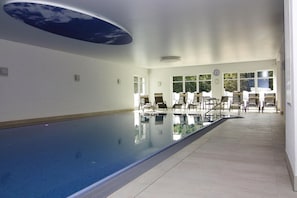 An indoor swimming pool with clear glass walls, a ceiling with a sky-like design, and a seating area with chairs and tables. - Apartments in the Avalon Hotel Bellevue, Fehmarn (Fehmarn)