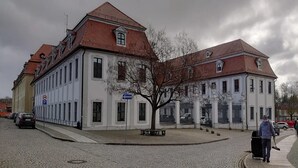 Ein historisches Gebäude mit einer symmetrischen Fassade, großen Fenstern und einem zentral platzierten Baum.