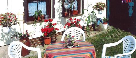 An outdoor patio with a table and chairs, surrounded by potted plants and a white wall with windows.