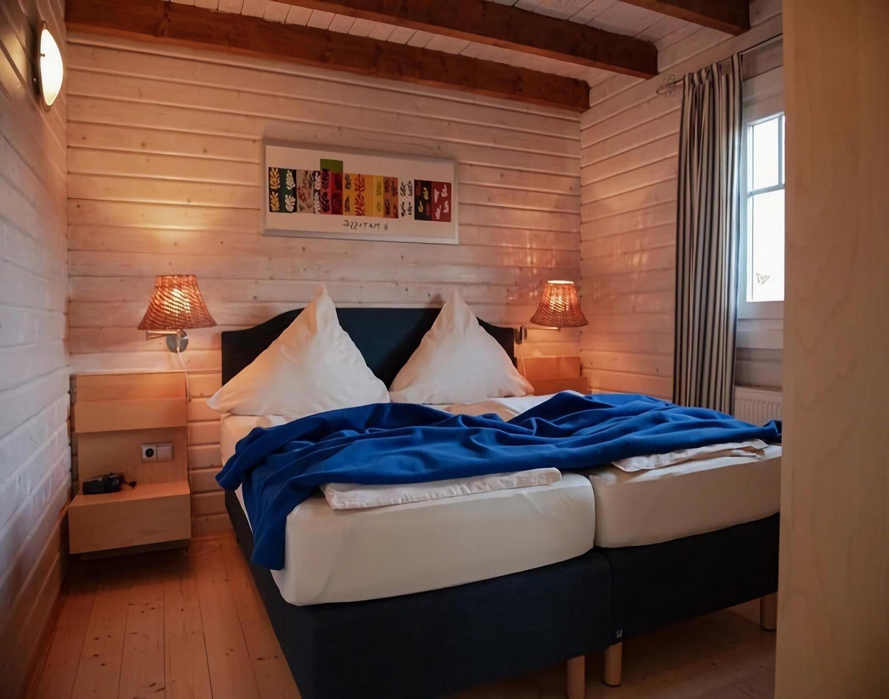 A cozy bedroom with a wooden headboard, two bedside lamps, a framed artwork above the bed, and a window with curtains.