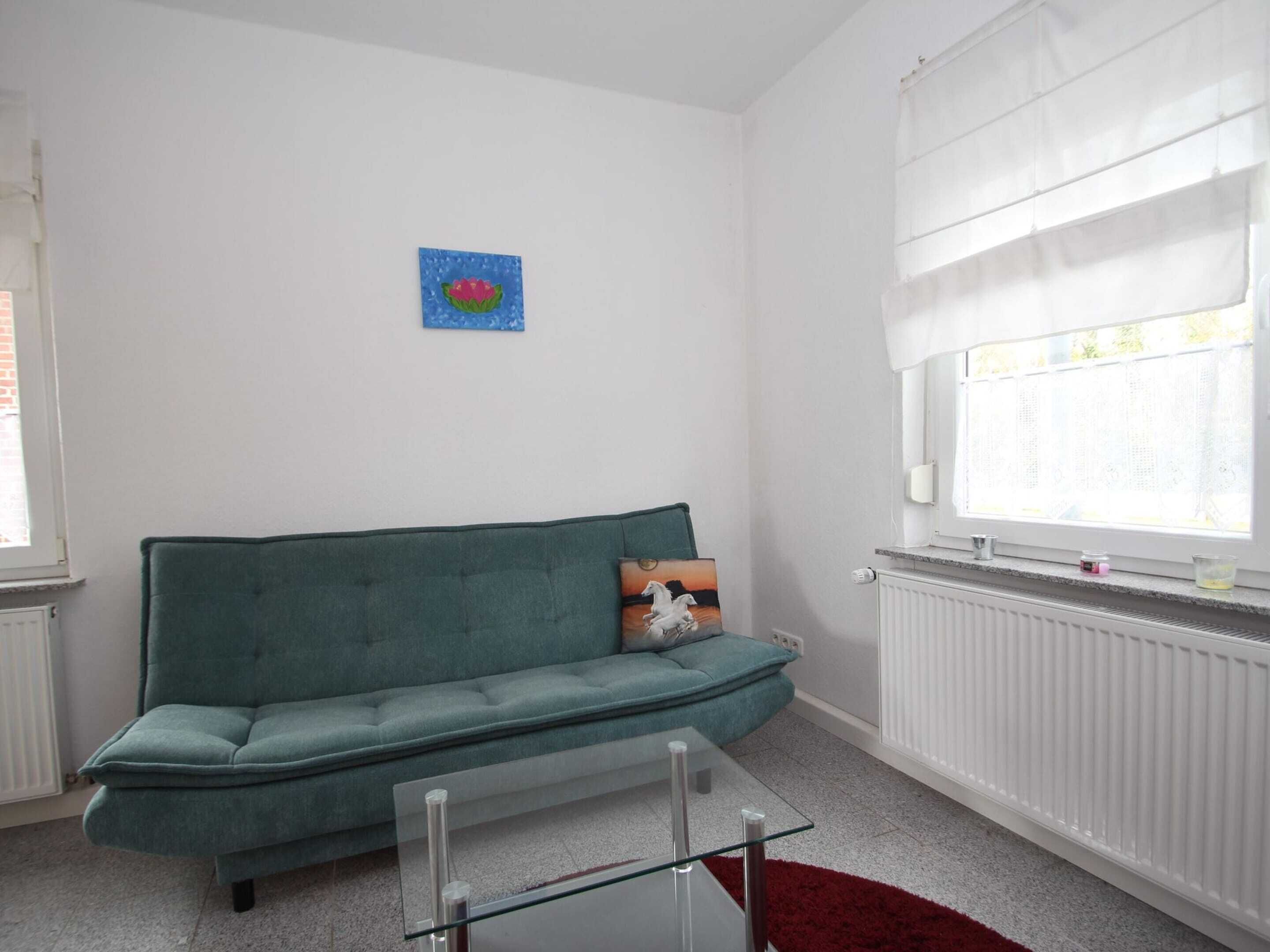 Un salon avec un canapé de couleur turquoise, une table basse en verre, un tapis rouge, une fenêtre avec des stores blancs et un petit tableau accroché au mur.