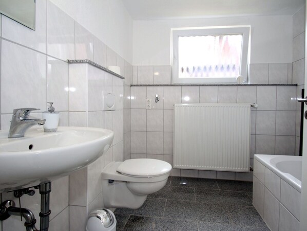 Une salle de bain avec un lavabo, des toilettes et une baignoire, tous de couleur blanche. Il y a une fenêtre au-dessus des toilettes et un radiateur au mur.