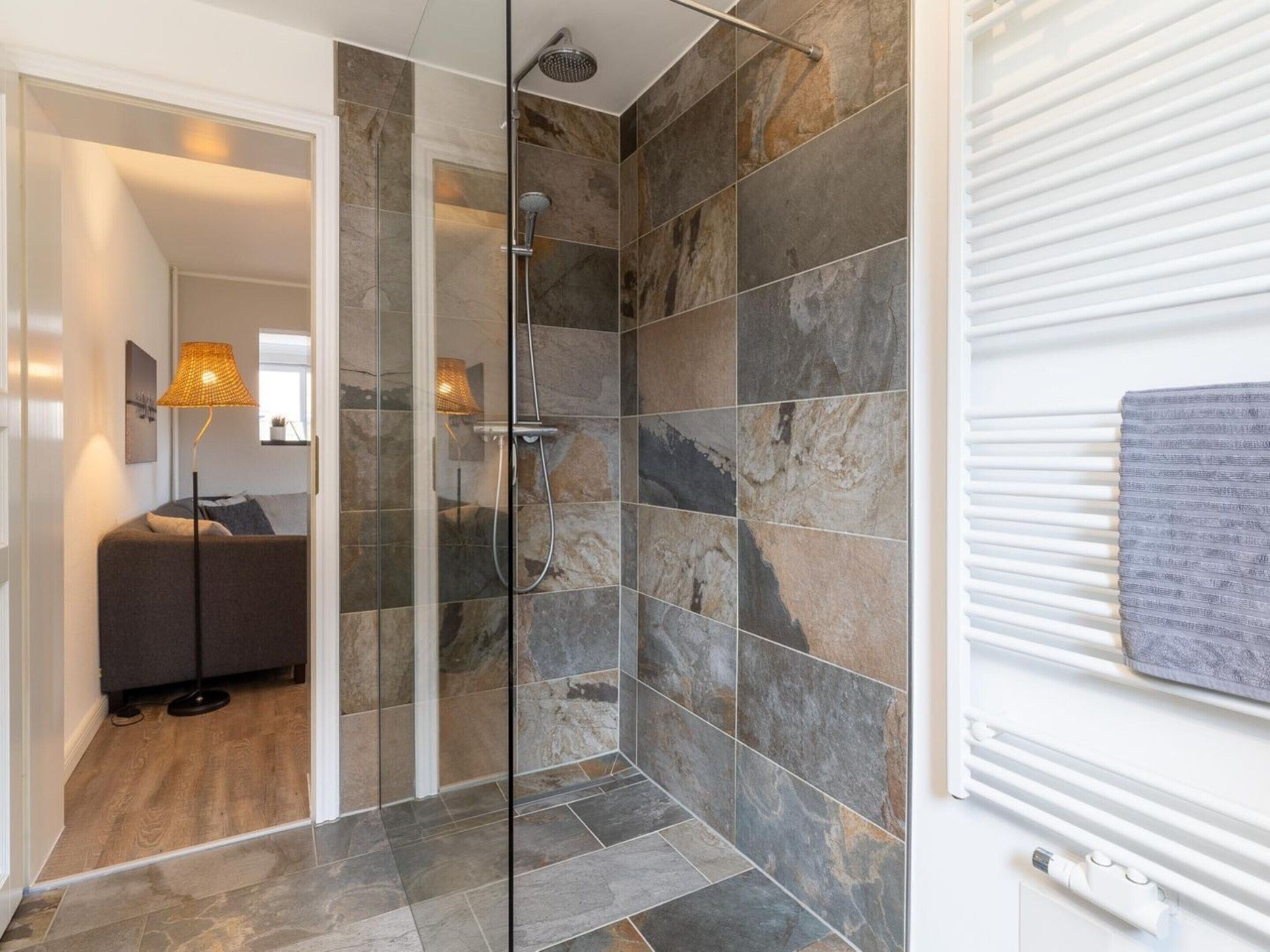 Un baño moderno con cabina de ducha de vidrio, baldosas de piedra y un sofá gris en la habitación contigua.