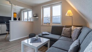 A modern living room with a grey sofa, a white coffee table, and a lamp. There is a kitchen area with a countertop and a hanging light fixture. - Holiday Dune Magic (St. Peter-Ording)