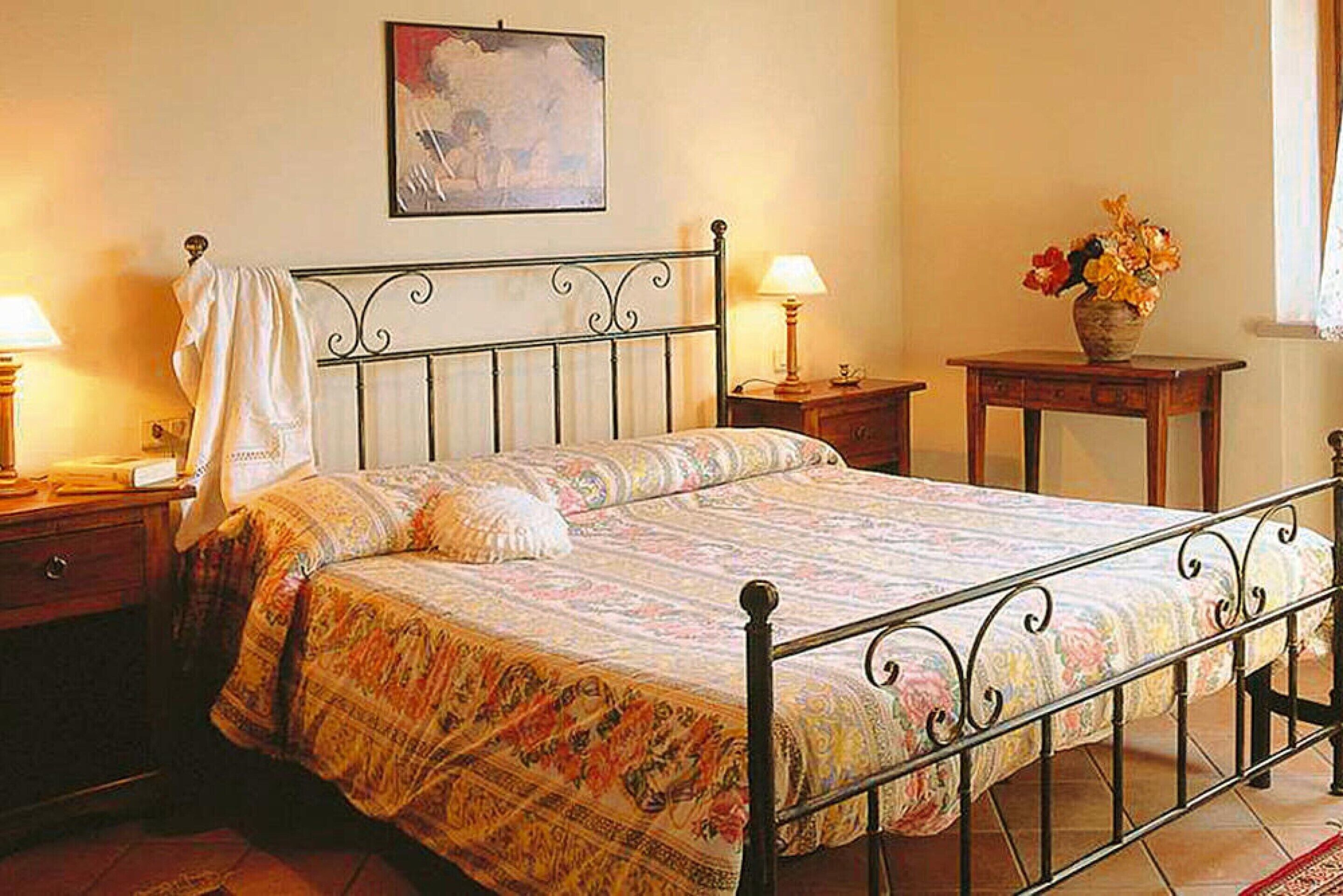 Una cama bien hecha con una colcha estampada, una mesita de noche con una lámpara y una pequeña mesa con un jarrón de flores.