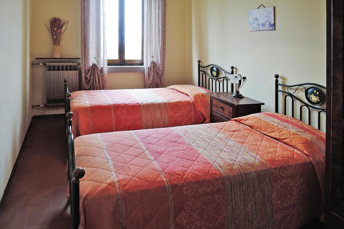A room with two single beds, a radiator, a window with curtains, a small table with a lamp, and a picture on the wall.