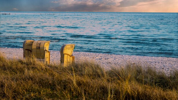 Ein Strand mit zwei gelben Stühlen, die zum Meer zeigen, einem sandigen Ufer und einem bewölkten Himmel.