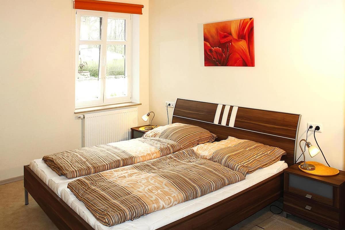 A neatly made bed with striped bedding, a window with a view, a bedside table with a lamp, and a painting on the wall.