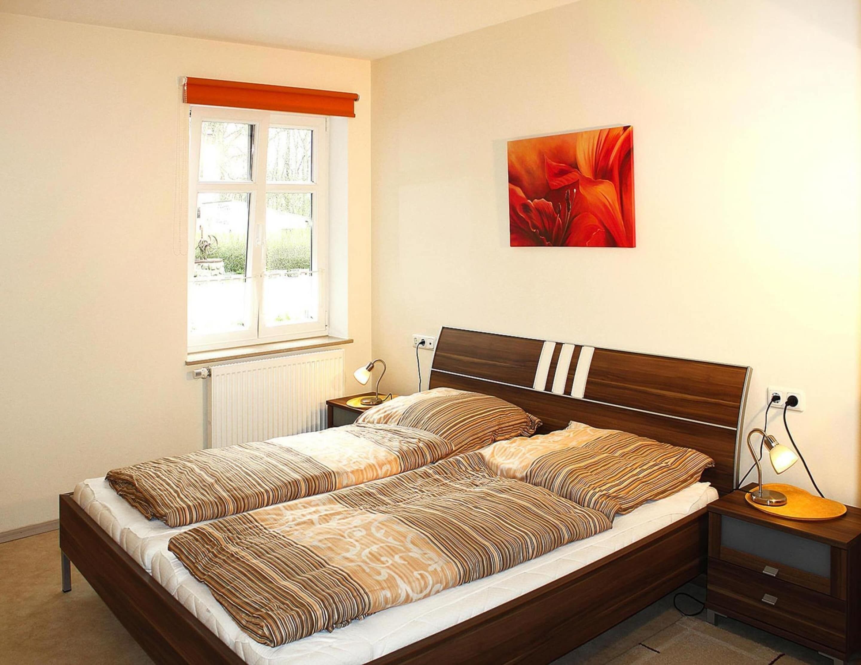 A neatly made bed with striped bedding, a window with a view, a bedside table with a lamp, and a painting on the wall.