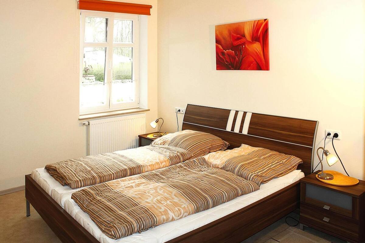 A neatly made bed with striped bedding, a window with a view, a bedside table with a lamp, and a painting on the wall.