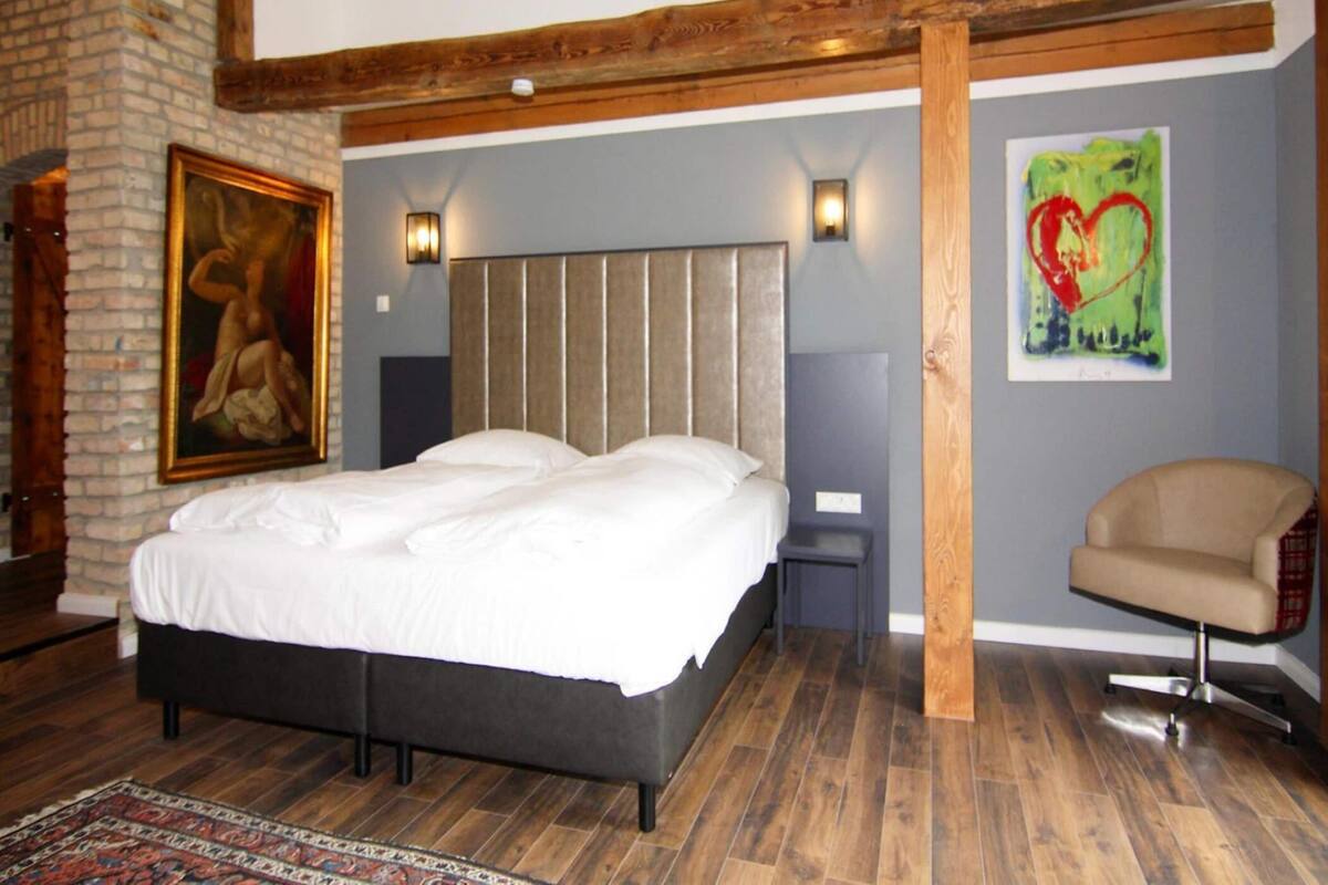 A bedroom with a large bed, a small table, a chair, a painting, and a wooden ceiling beam.