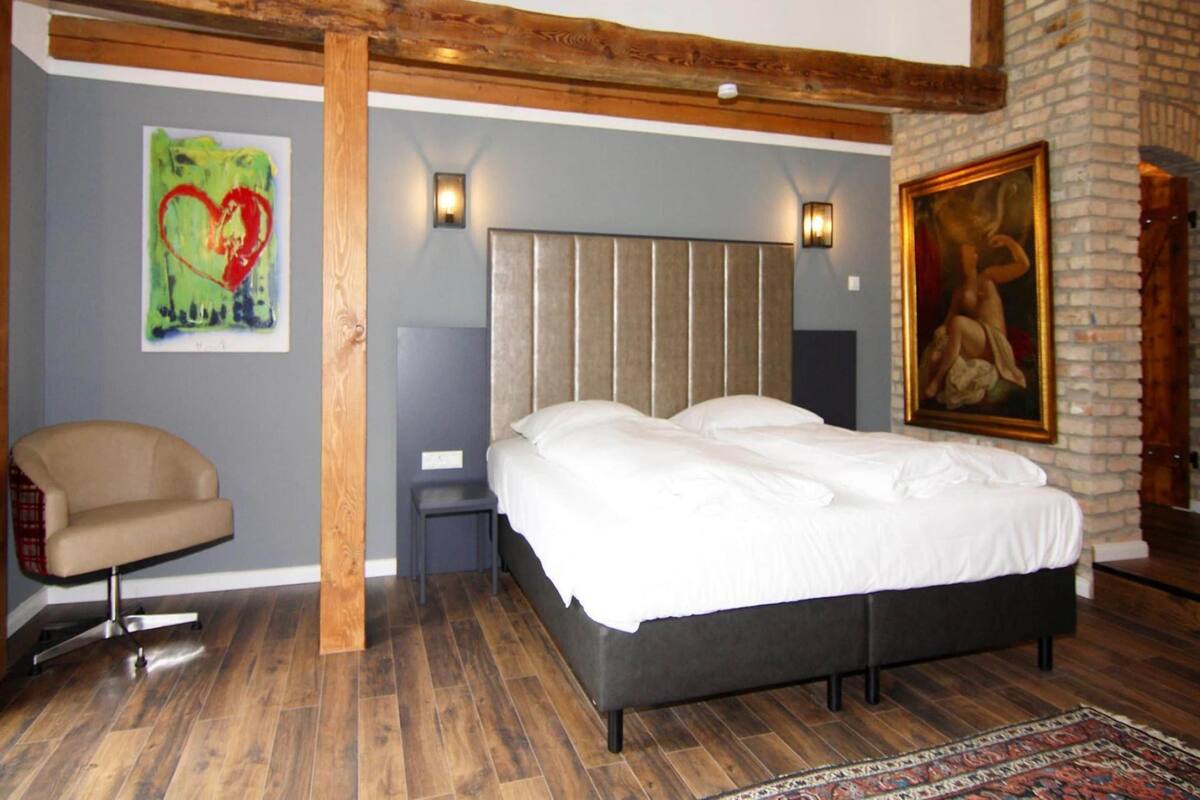 A bedroom with a large bed, a chair, a painting, and a fireplace.