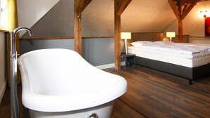 A bedroom with a sloped ceiling, wooden beams, a white bathtub, a bed with white bedding, and two bedside lamps. - Holiday Home Birgit, Wendorf (Kuhlen-Wendorf)