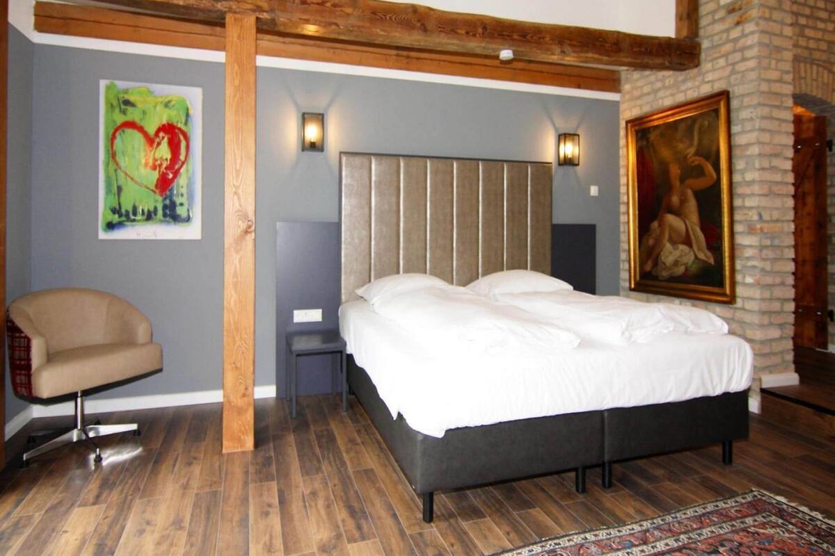 A bedroom with a bed, a chair, a painting, and a fireplace.