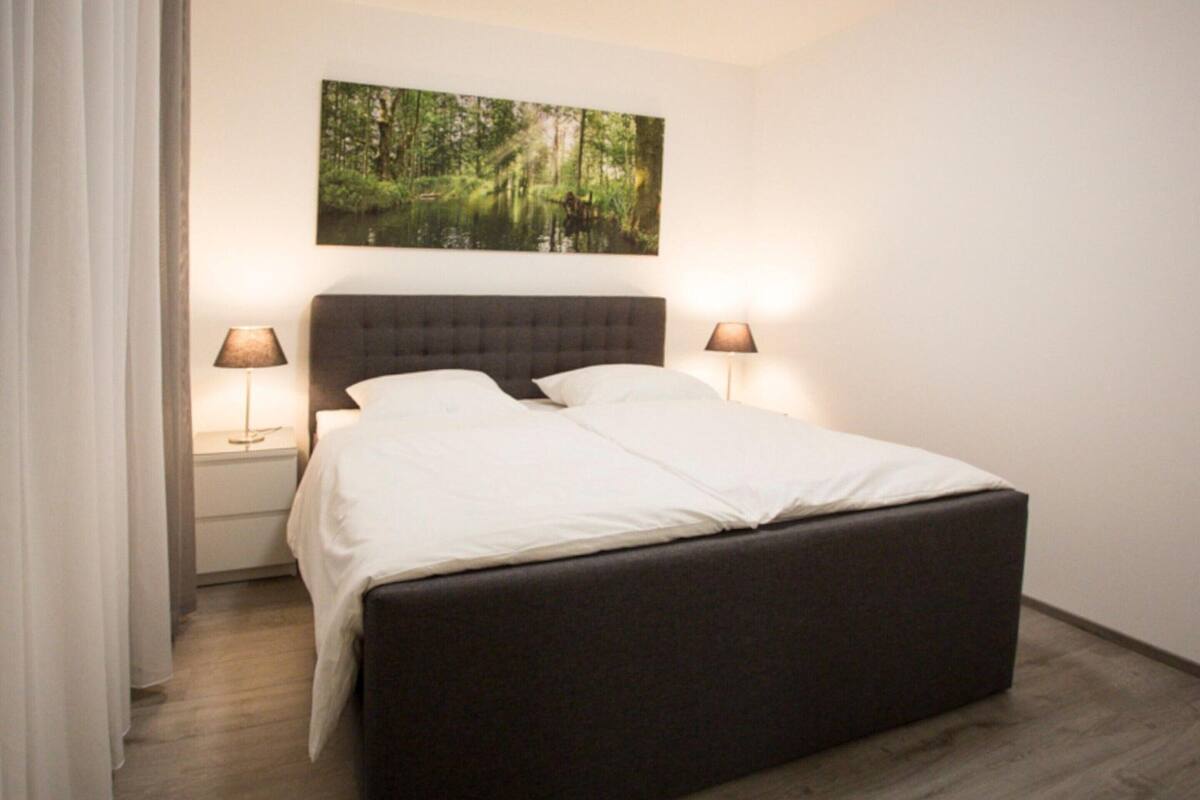 A modern bedroom with a large bed, two bedside tables with lamps, and a forest-themed painting on the wall.