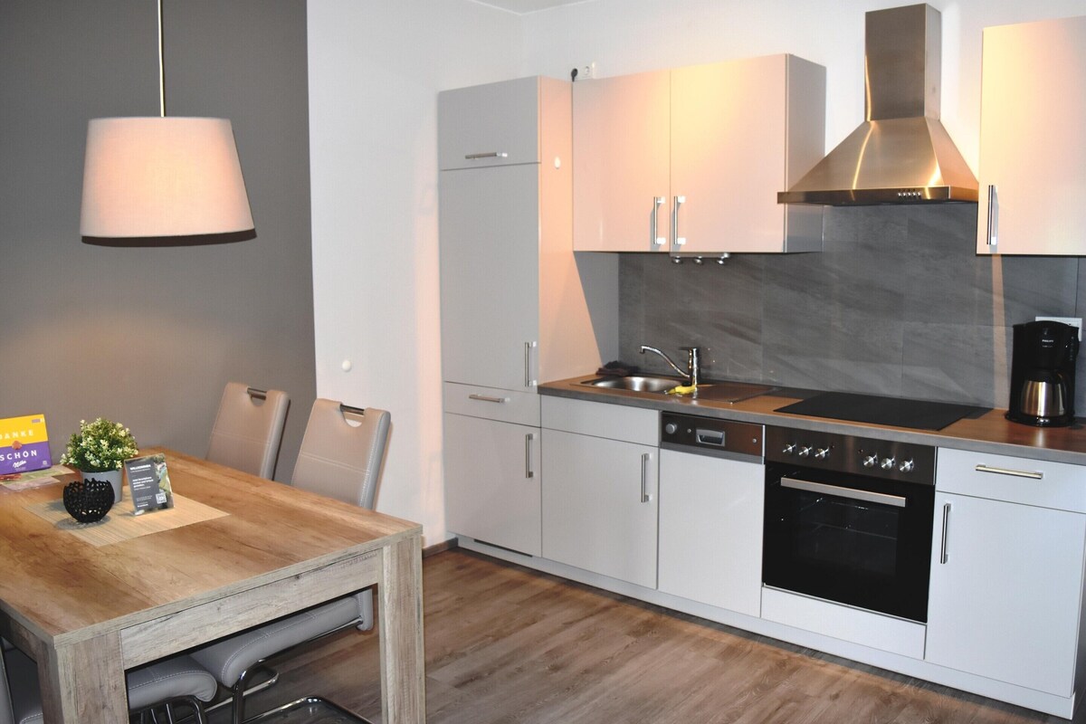 A modern kitchen with white cabinets, a stainless steel range hood, and a wooden dining table.