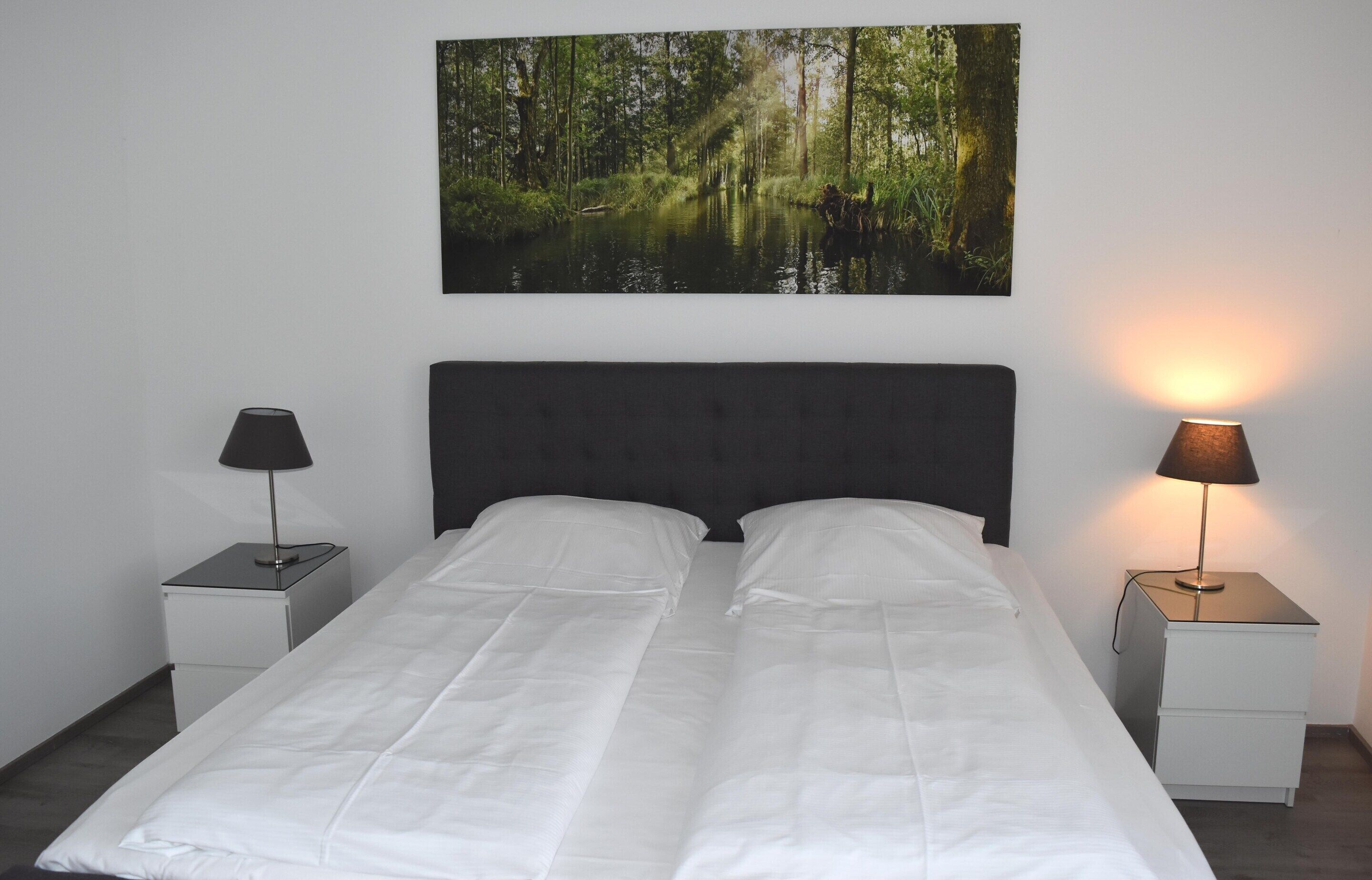 A bed with white linens, two bedside tables with lamps, and a forest-themed wall hanging.