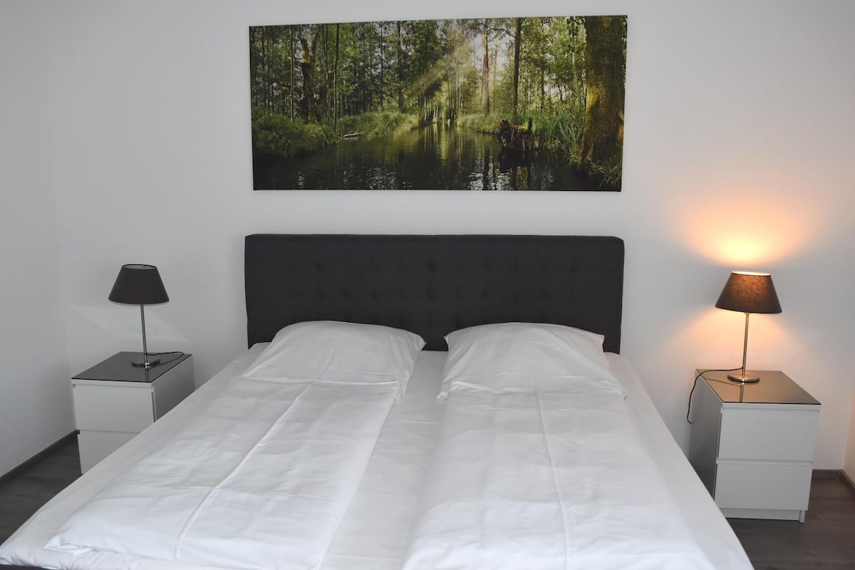 A bed with white linens, two bedside tables with lamps, and a forest-themed wall hanging.