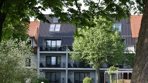 Ein mehrstöckiges Wohngebäude mit Balkonen, einem Rasen vor der Fassade und einem Baum im Vordergrund.