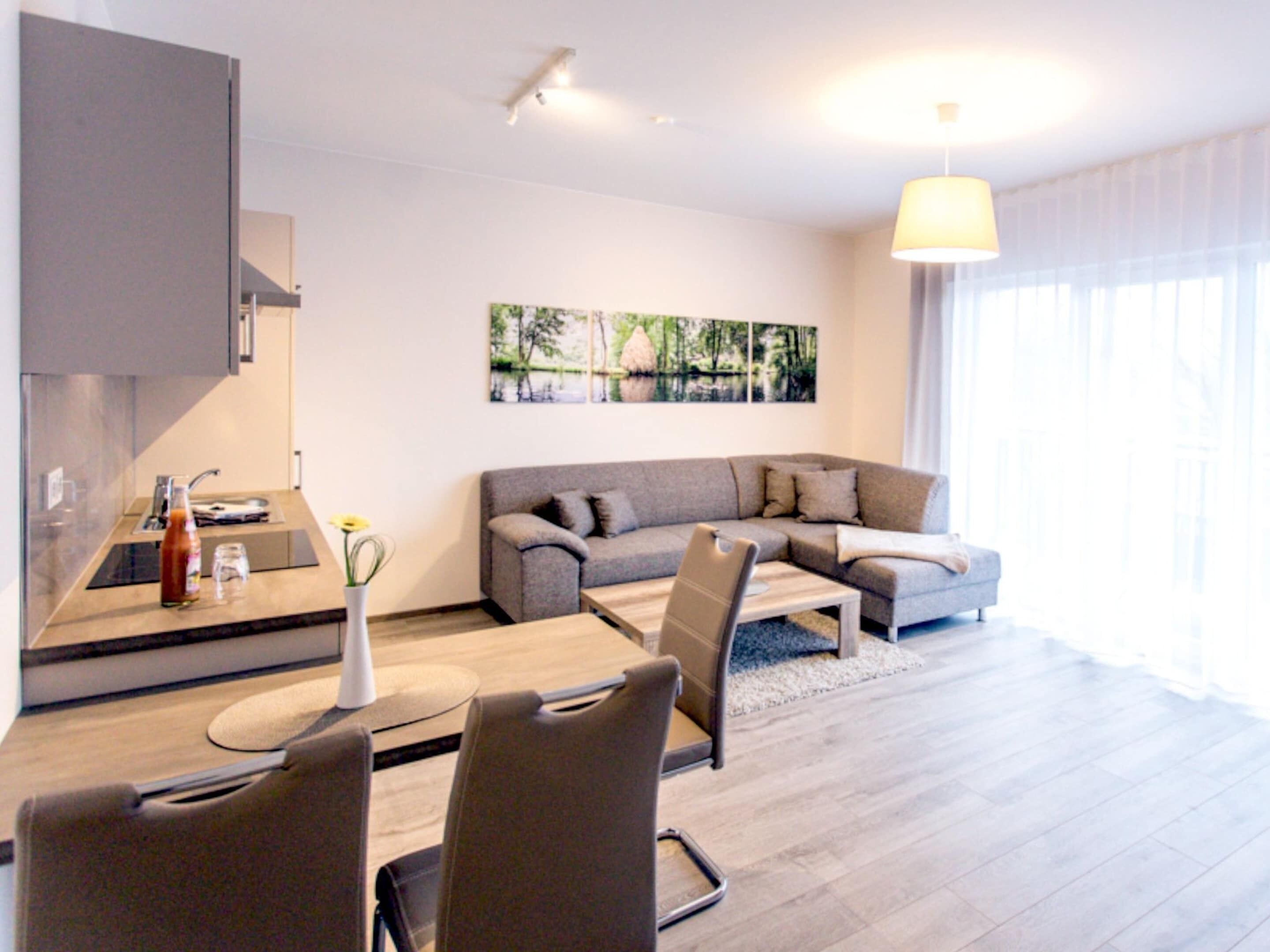 Ein modernes Wohnzimmer mit einem grauen Sofa, einem Esstisch mit Stühlen, einer Küchenzeile und einem großen Fenster mit Vorhängen.