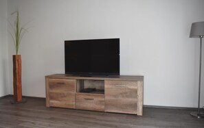 A wooden TV stand with a flat-screen TV, a small cabinet, and a tall floor lamp against a plain wall. - Apartment in Lubben Near the Water (Lübben (Spreewald))