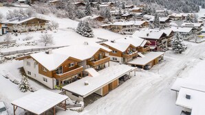 Un quartier résidentiel enneigé, avec de nombreuses maisons, dont une grande maison en bois dotée d’un balcon et d’un espace extérieur aménagé et abrité.