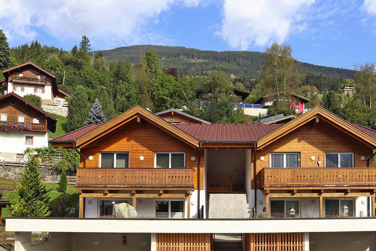 A wooden chalet with a balcony, set against a mountainous backdrop.