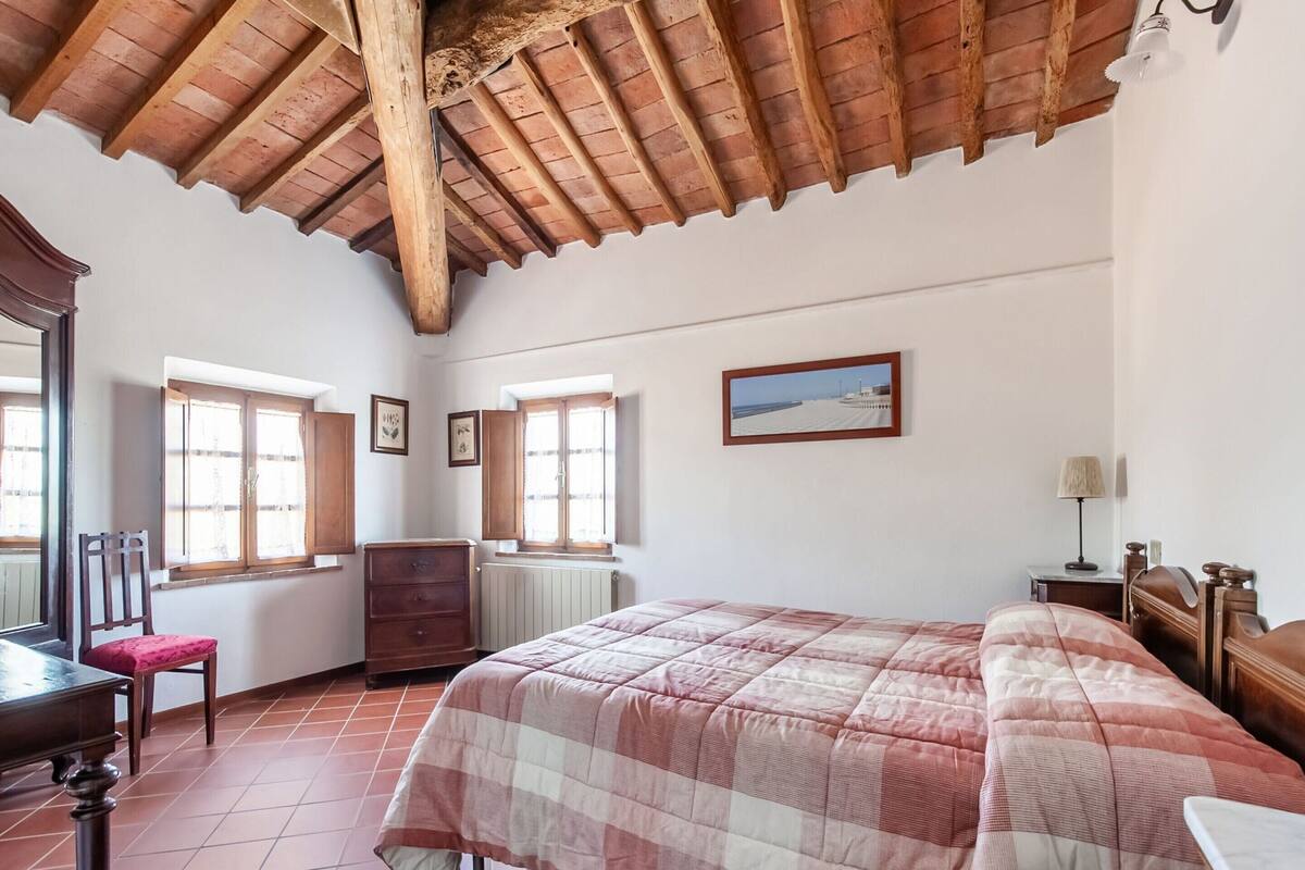 A bedroom with a wooden ceiling, a bed with a checkered bedspread, a dresser, a chair, and a window with a view of the sea.