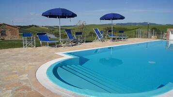 A swimming pool with blue chairs and umbrellas, surrounded by stone tiles and a view of green hills.