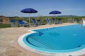 Piscina con sillas y sombrillas azules, rodeada de baldosas de piedra y vistas a colinas verdes.