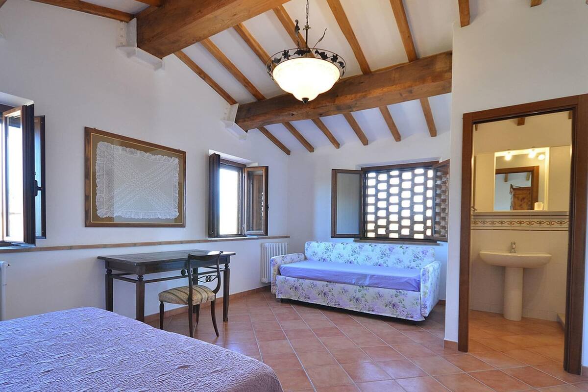 A bedroom with a bed, a table, a chair, a sofa, a window, a radiator, and a bathroom with a sink and mirror.