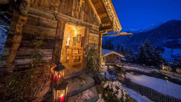 Eine Holzhütte mit einer Holztreppe, die zum Eingang führt, einer beleuchteten Statue im Inneren und einer verschneiten Landschaft mit Bergen im Hintergrund.