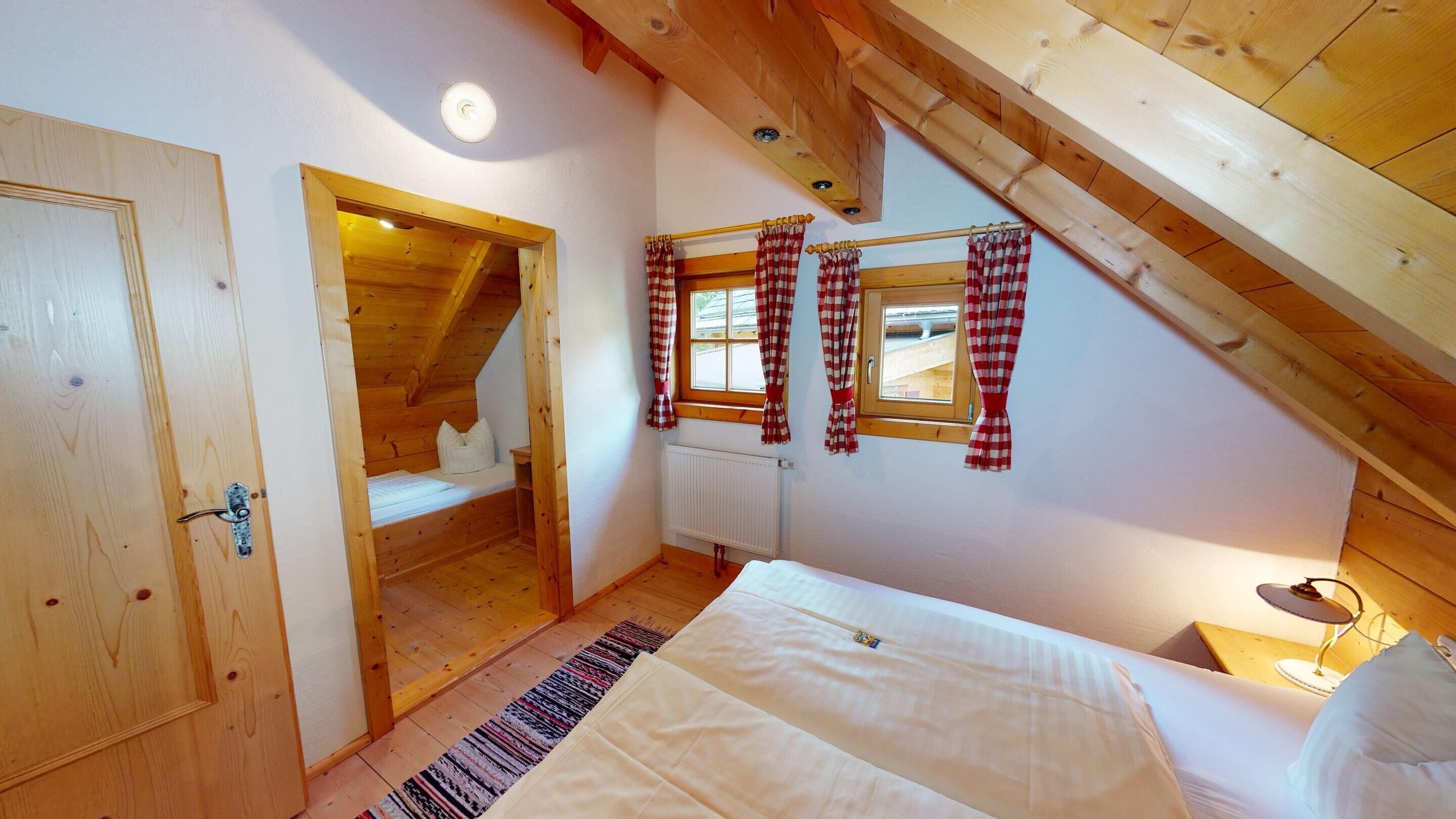 Een slaapkamer met een houten plafond, een bed, een raam met geruite gordijnen en een deur.
