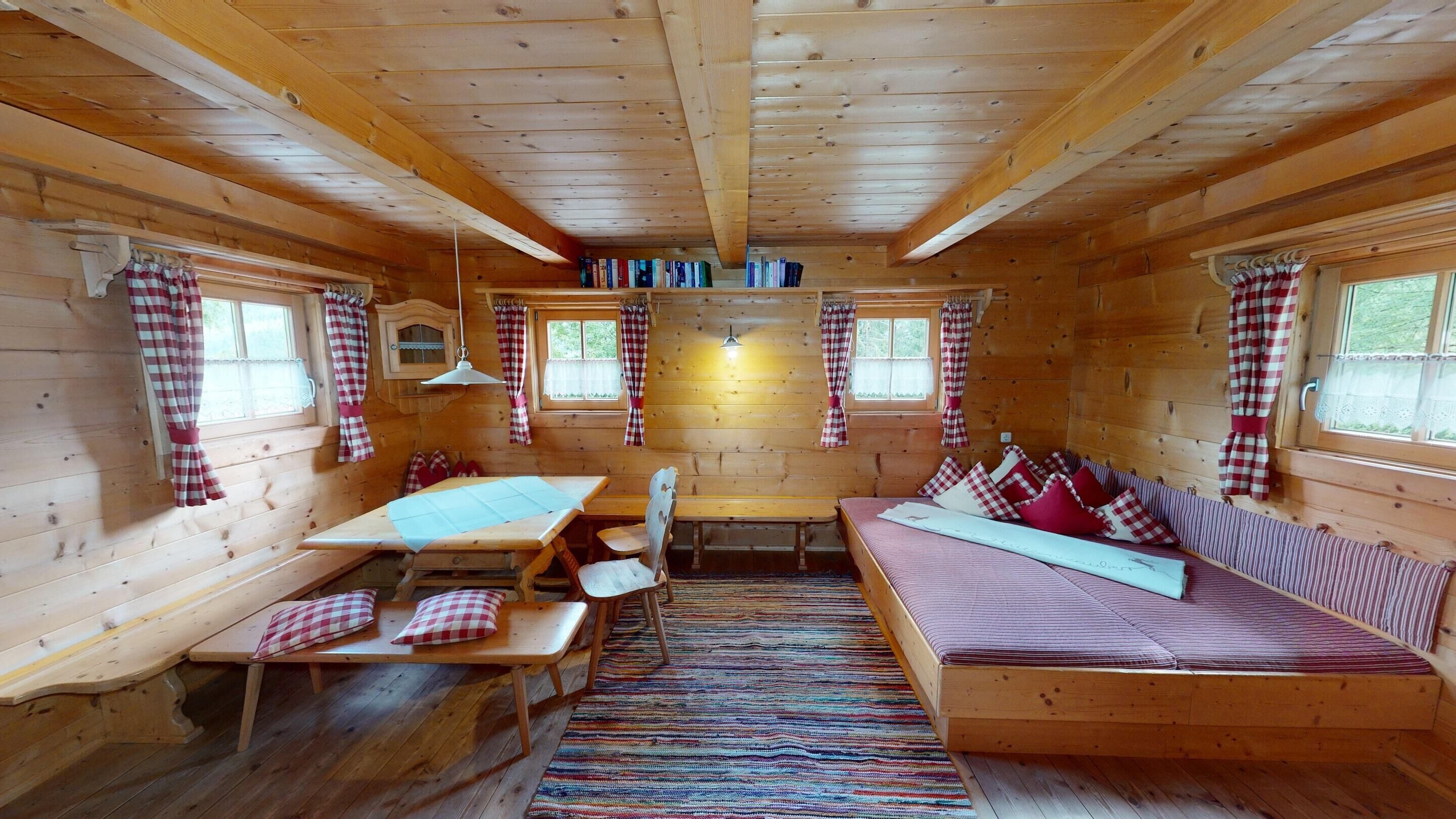 Een gezellig chalet binnenin, met een houten vloer, een eettafel, bankjes, een bank en een boekenkast.