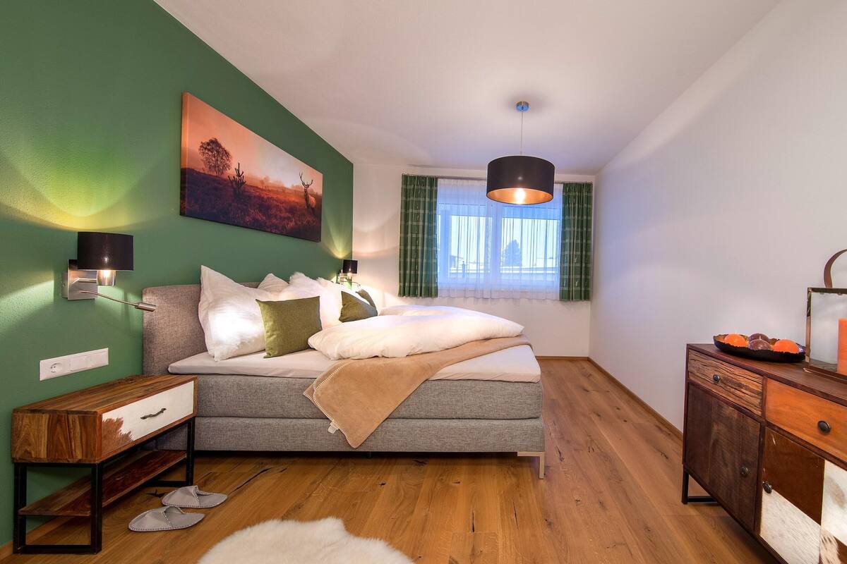 A bedroom with a bed, bedside tables, a dresser, a window with curtains, and a painting on the wall.
