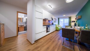 Eine moderne Küche mit weißen Schränken, einer Holz Arbeitsplatte und integrierten Geräten. Es gibt eine Essecke mit Tisch und Stühlen sowie einen Ausblick ins Freie durch große Fenster.