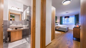 Una habitación de hotel moderna con piso de madera, una cama grande, una mesita de noche y un baño con ducha y lavamanos.