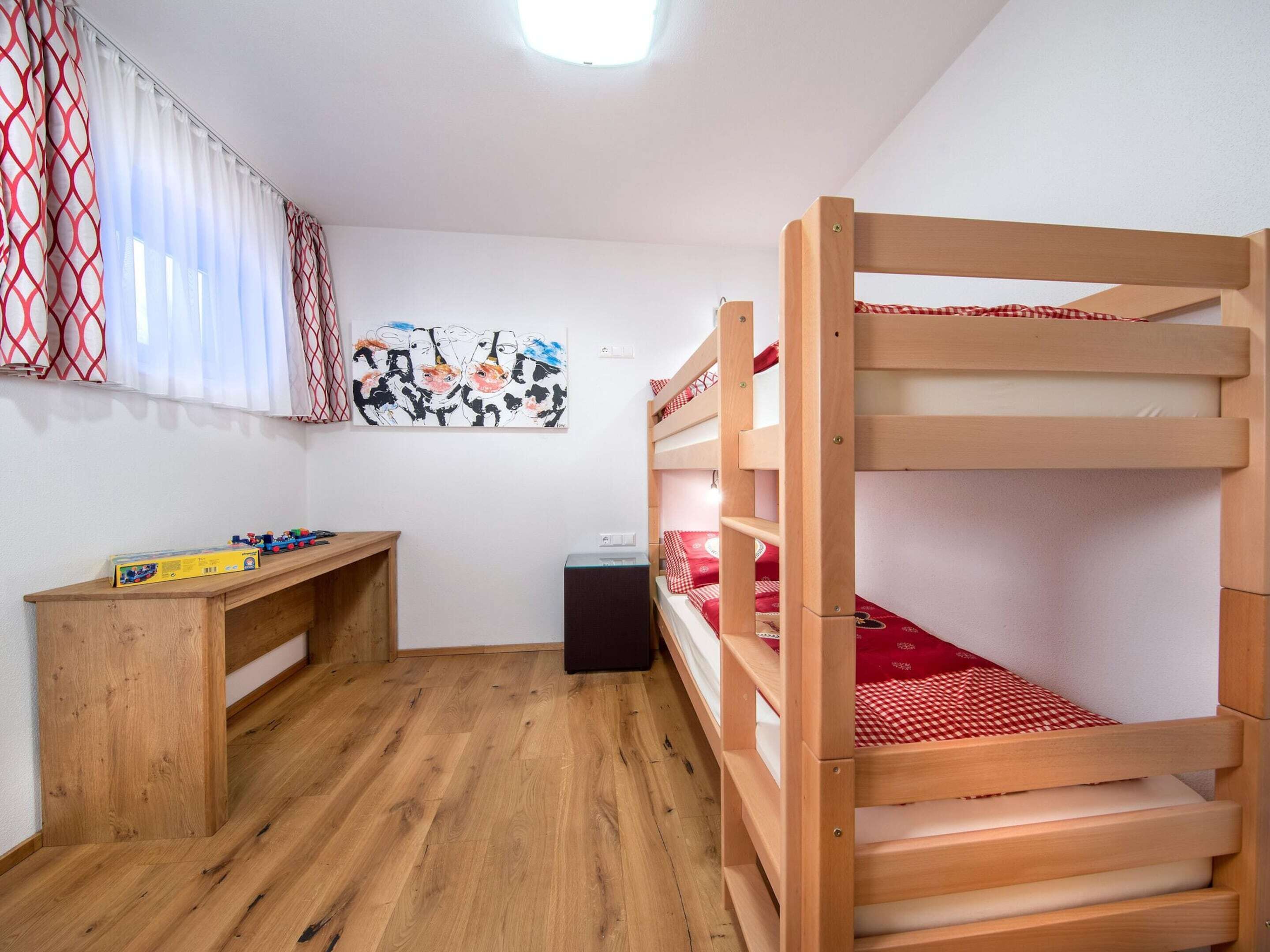 A bunk bed room with a wooden desk and a small table, a painting on the wall, and a window with curtains.