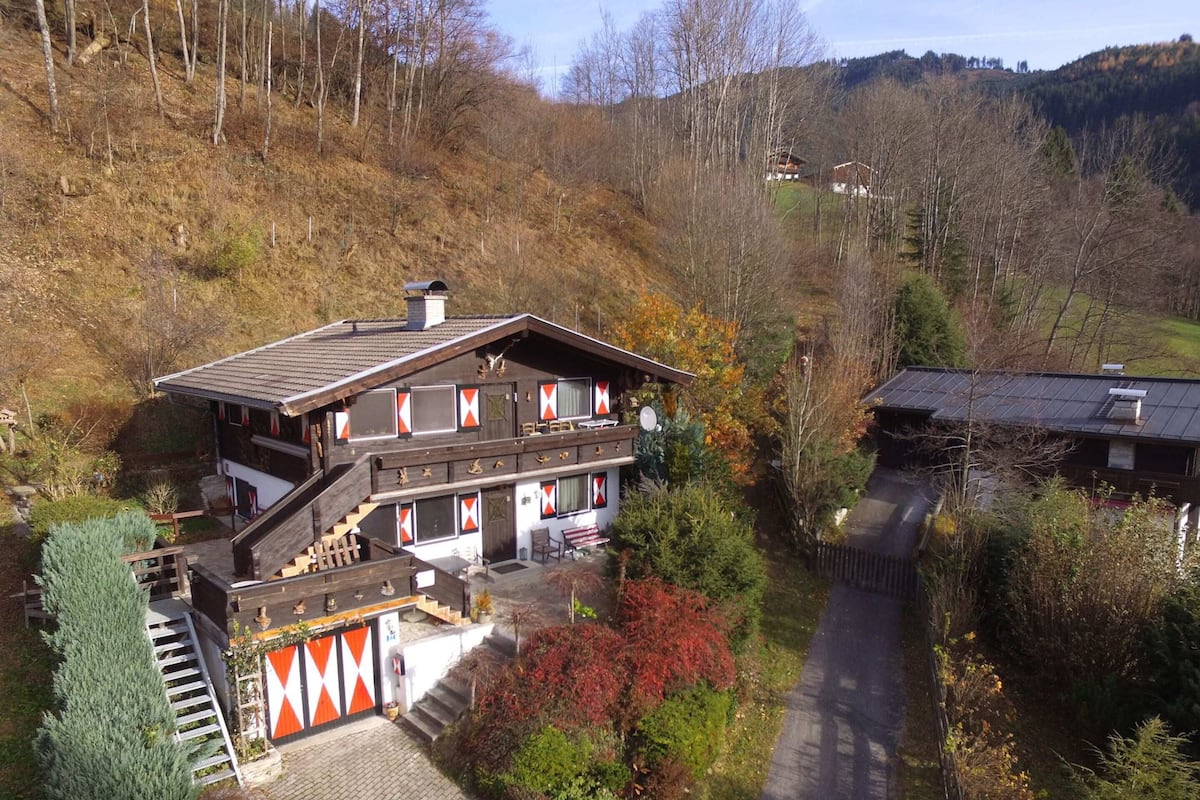 A two-story chalet with a steep roof, a wooden staircase, and a view of the surrounding forest and hills.