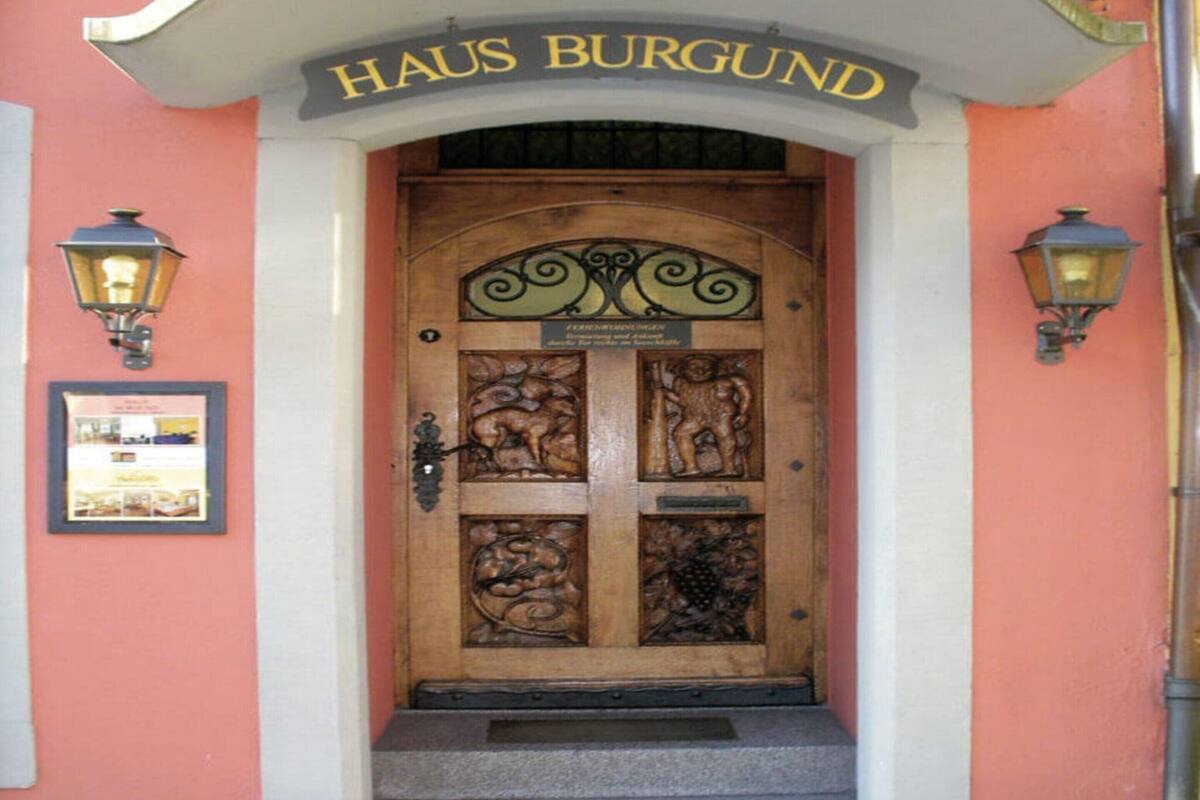 Eine Holztür mit aufwendigen Schnitzereien, ein Schild mit der Aufschrift „HAUS BURGUND“ darüber und je eine Wandleuchte auf beiden Seiten.