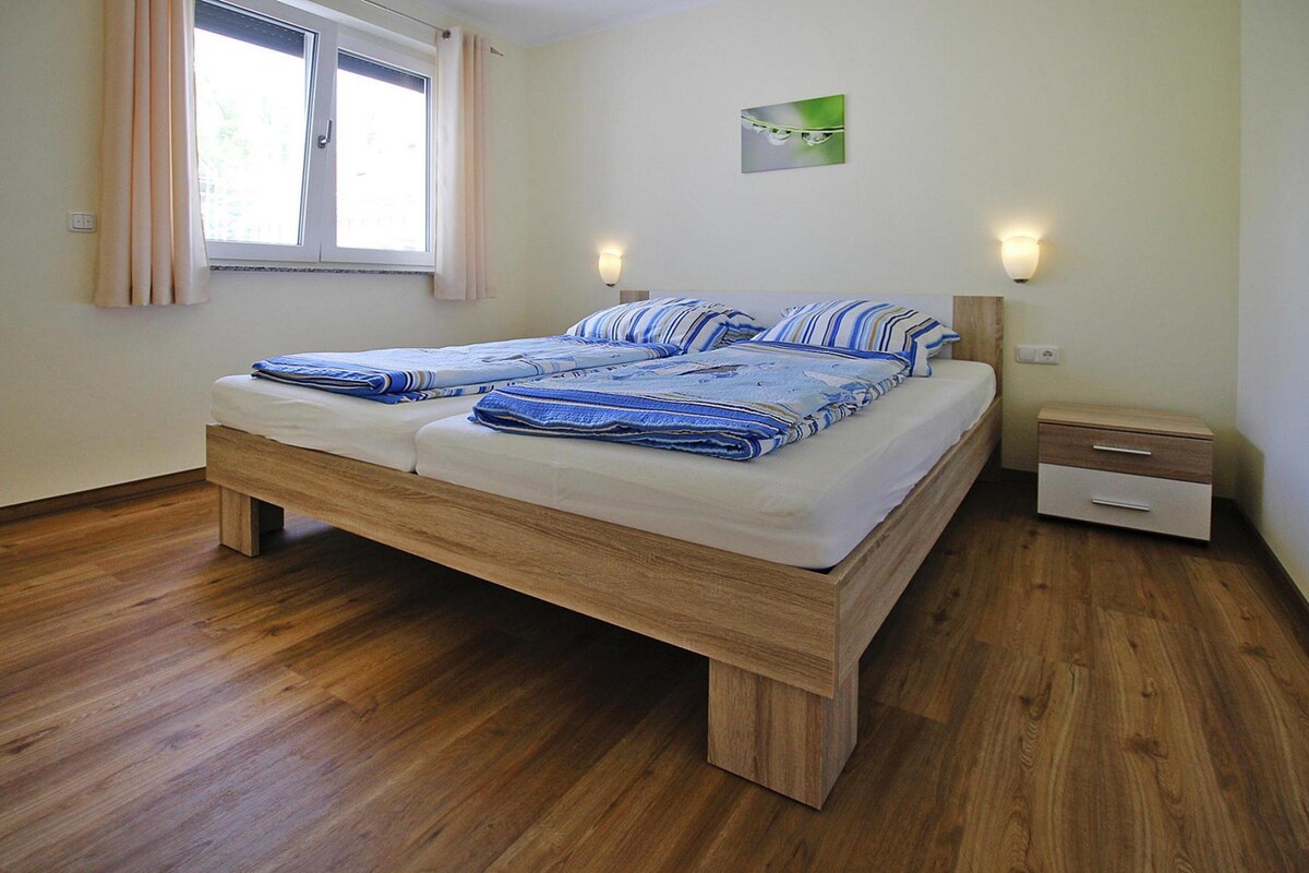 A double bed with a wooden frame, two bedside tables with lamps, a window with curtains, and a small wall-mounted picture.