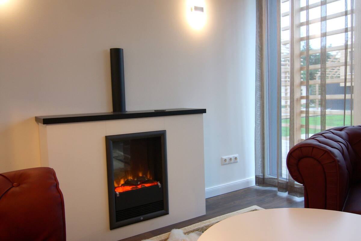 A modern living room with a fireplace, a red leather sofa, and a view of a green outdoor area through large windows.